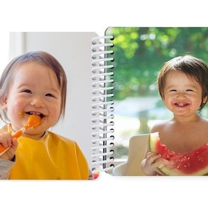 May include: Two babies, one smiling while eating with a spoon, the other smiling while eating a slice of watermelon.