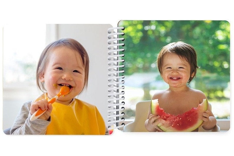 May include: Two babies, one smiling while eating with a spoon, the other smiling while eating a slice of watermelon.
