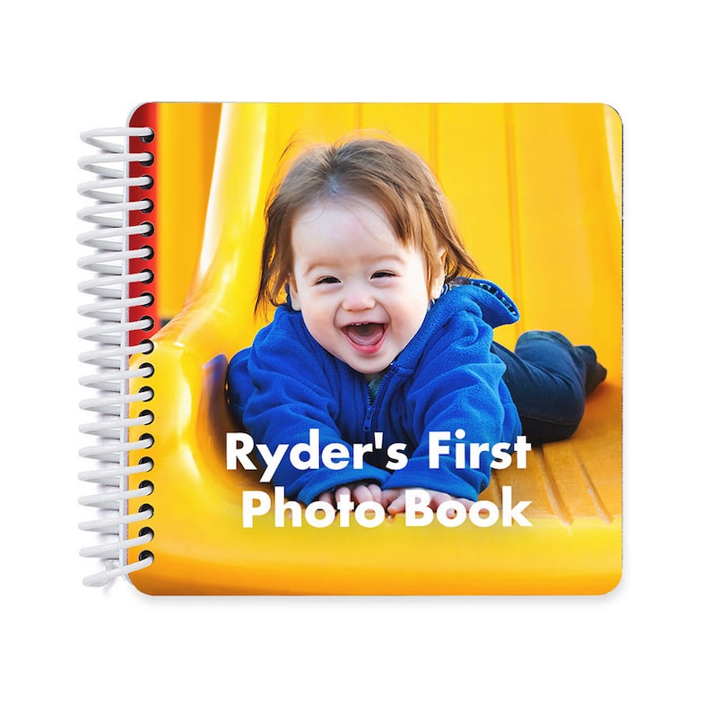 May include: A yellow photo book with a white spiral binding. The cover features a photo of a baby boy laughing on a yellow slide. The text "Ryder's First Photo Book" is printed in white on the cover.