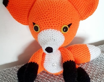 Puede incluir: Un juguete de peluche de zorro naranja tejido a crochet con orejas, patas y nariz negras. El zorro tiene grandes ojos amarillos y un hocico blanco.