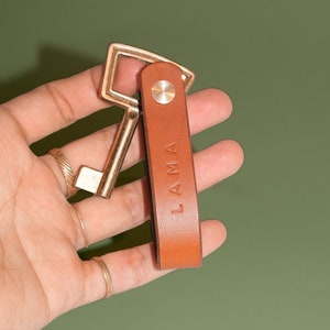 May include: A brown leather keychain with the word "LAMA" embossed on it. The keychain has a gold metal key ring attached to it.