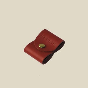 Puede incluir: Pequeño estuche de cuero rojo con solapa asegurada por un broche de color latón. La palabra "BERLIN" está grabada en relieve en la solapa. El estuche está diseñado para guardar objetos pequeños.