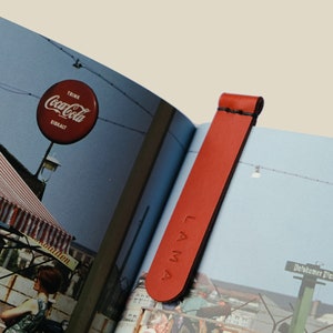 Può includere: Un segnalibro in pelle rossa con la scritta "LAMA" in rilievo, inserito in un libro. Il segnalibro è di forma rettangolare con la parte superiore arrotondata. Le pagine del libro mostrano un'insegna vintage della Coca-Cola.