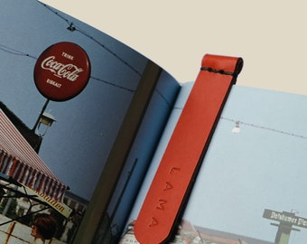 May include: A red leather bookmark with the word "LAMA" embossed on it, inserted into a book. The bookmark is a solid, rectangular shape with a rounded top. The book pages show a vintage Coca-Cola sign.