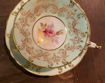 May include: A vintage teacup and saucer set. The cup and saucer are light green with gold filigree and a pink rose in the center. The rims are scalloped and trimmed with gold. The handle of the cup is also gold.