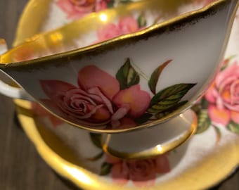May include: White teacup and saucer set with pink rose floral design and gold trim. The teacup has a delicate handle and sits on a matching saucer. The set is likely vintage or antique.