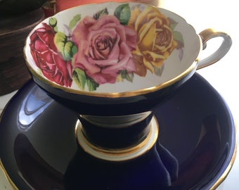 May include: A vintage teacup and saucer set. The teacup features a floral design with red, pink, and yellow roses. The cup and saucer are a deep blue with gold trim. The set is designed for tea parties and special occasions.