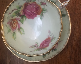 May include: A vintage teacup and saucer set. The cup and saucer are light green with a gold trim and feature a floral design of pink and orange roses with green leaves. The set is placed on a wooden surface.