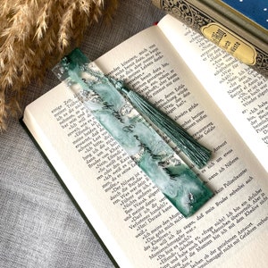 Könnte beinhalten: Handgefertigter Lesezeichen mit türkis-weißem Harzdesign, Silberflocken und passendem Quasten. Der Lesezeichen hat den Text "a cup of tea & a good book". Es liegt auf einem offenen Buch.