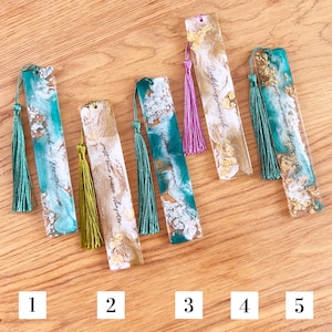 Könnte beinhalten: Fünf Lesezeichen aus Kunstharz mit Quasten. Die Lesezeichen weisen Designs in Türkis, Gold und Weiß mit Text auf. Jedes Lesezeichen hat eine andersfarbige Quaste, darunter Grün, Gold und Lila. Die Lesezeichen sind rechteckig und haben eine glänzende Oberfläche.