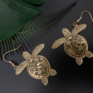 Puede incluir: Un par de pendientes dorados con forma de tortuga. Cada pendiente presenta un diseño detallado de tortuga con patrones grabados y detalles florales. Los pendientes tienen un cierre de gancho y se exhiben sobre una superficie negra con una hoja verde.