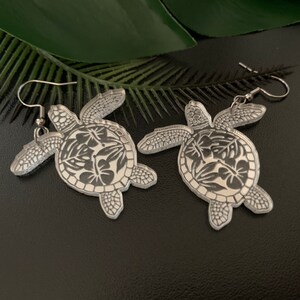 Puede incluir: Un par de pendientes plateados con forma de tortuga. Cada pendiente presenta una tortuga con un diseño floral detallado en el caparazón. Los pendientes tienen un cierre de gancho y se exhiben sobre una superficie negra con una hoja verde.