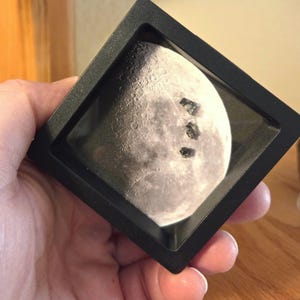 Può includere: Una scatola nera quadrata che mostra un'immagine dettagliata della superficie lunare. Tre piccoli oggetti scuri sono posizionati sulla superficie lunare. La scatola è tenuta nella mano di una persona.