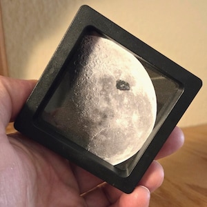 Peut inclure: Un cadre carré noir contenant une image détaillée de la surface lunaire, avec des cratères et des textures visibles. Un objet sombre de forme irrégulière est visible sur la lune. Le cadre est tenu dans une main.