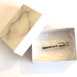 May include: A small glass vial pendant with a silver-toned metal cap and loop, containing tiny dark grey mineral fragments. The pendant is resting inside a white box with a marble-patterned lid, lined with clear bubble wrap for protection.
