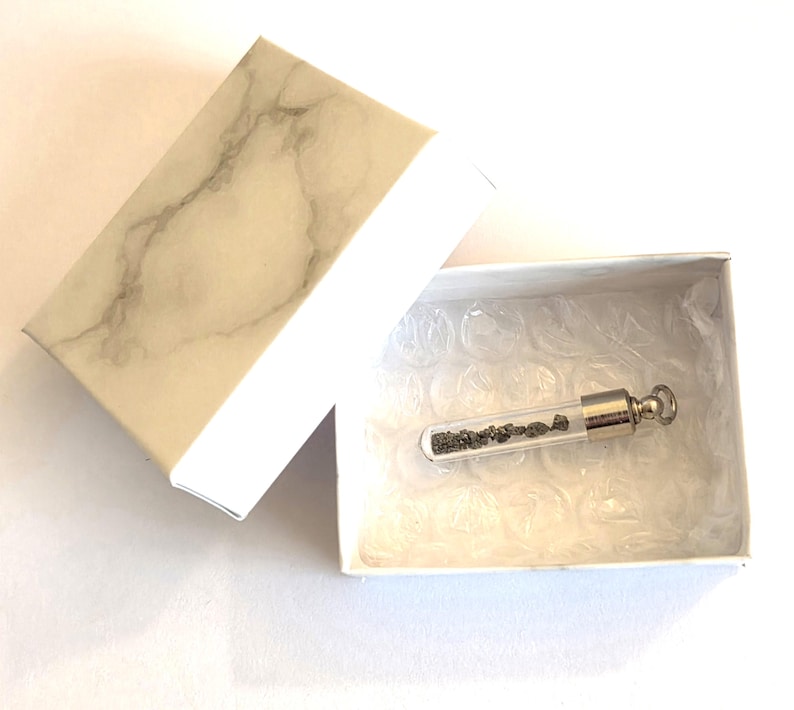 May include: A small glass vial pendant with a silver-toned metal cap and loop, containing tiny dark grey mineral fragments. The pendant is resting inside a white box with a marble-patterned lid, lined with clear bubble wrap for protection.