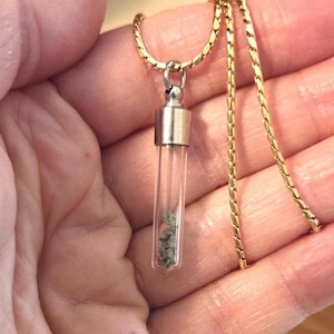 May include: A gold-toned chain necklace featuring a clear glass vial pendant. The cylindrical vial is capped with a metallic silver-colored top and contains small, grey, textured mineral fragments visible through the transparent glass walls.