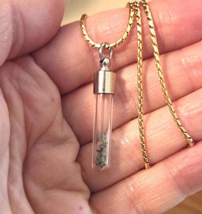 May include: A gold-toned chain necklace featuring a clear glass vial pendant. The cylindrical vial is capped with a metallic silver-colored top and contains small, grey, textured mineral fragments visible through the transparent glass walls.
