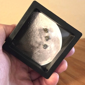 Puede incluir: Un marco de exhibición cuadrado negro que contiene un inserto circular con una impresión fotográfica detallada de la superficie lunar. Tres pequeños cristales de pirita metálicos y cúbicos están colocados en fila sobre la imagen.