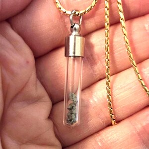 May include: A clear glass vial pendant suspended from a gold-toned chain. The cylindrical vial features a metallic silver-colored cap and contains small, grey, textured mineral fragments visible through the transparent glass walls.