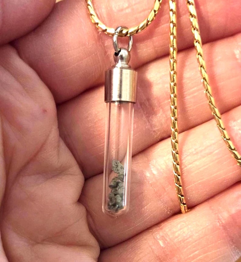 May include: A clear glass vial pendant suspended from a gold-toned chain. The cylindrical vial features a metallic silver-colored cap and contains small, grey, textured mineral fragments visible through the transparent glass walls.