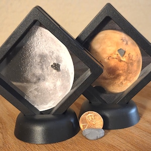 May include: Two square black floating display frames containing circular images of the Moon and Mars, each mounted on a black circular base. A copper-colored US penny is positioned between the two stands for scale, next to a small gray piece of clay.
