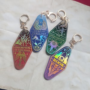 May include: Four keychains with gold-colored key rings. Each keychain is a different color: red, blue, green, and purple. Each keychain has a different design and text: "Champion of Kirkwall", "Hero of Ferelden", "The Inquisitor", and "Rook".
