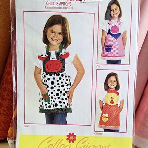 May include: A sewing pattern envelope titled Farm Friends Child's Aprons, sizes 3-8, by Cotton Ginnys. The cover features three aprons designed as a cow, a pig, and a chicken, each with matching pockets and animal-themed appliques.