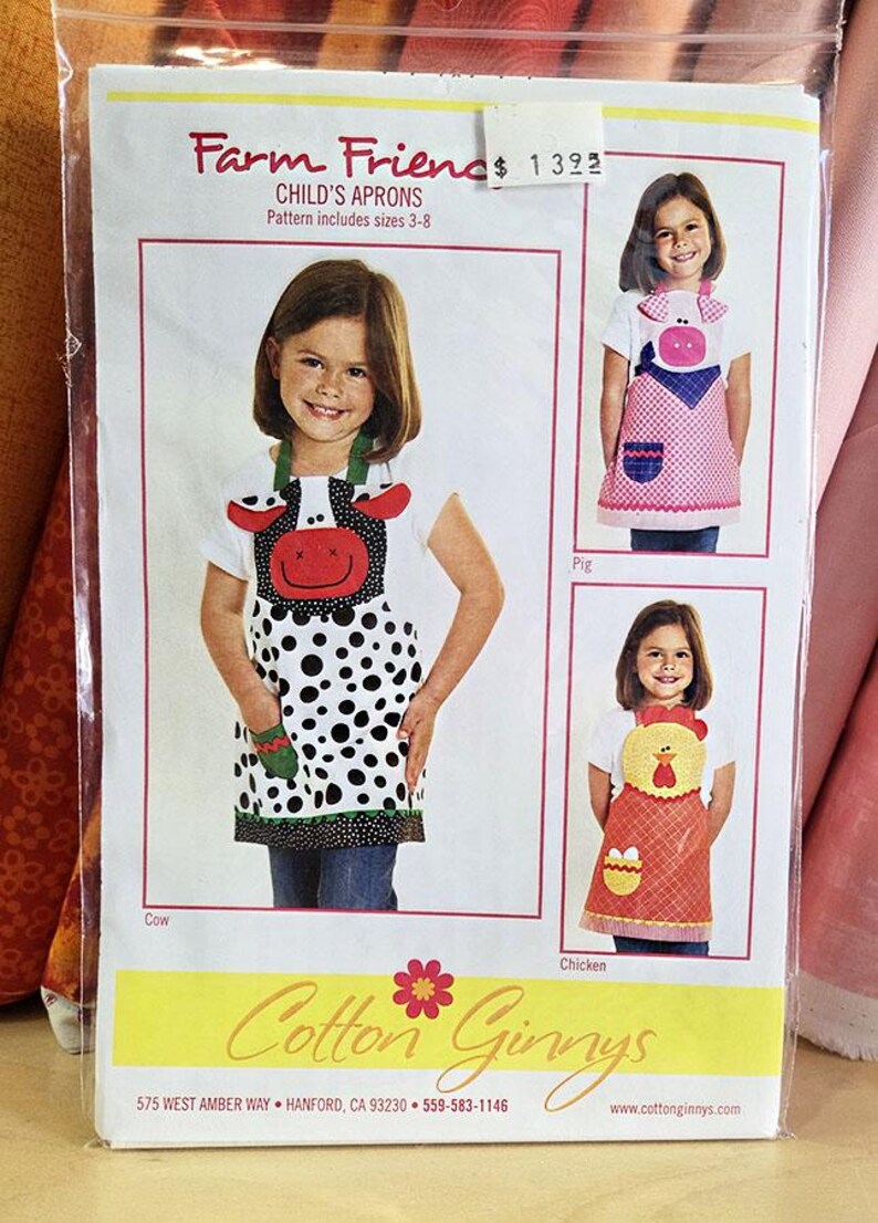 May include: A sewing pattern envelope titled Farm Friends Child's Aprons, sizes 3-8, by Cotton Ginnys. The cover features three aprons designed as a cow, a pig, and a chicken, each with matching pockets and animal-themed appliques.