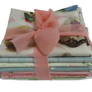 May include: A stack of fabric squares featuring illustrations from the classic children's book "The Tale of Peter Rabbit". The fabric squares are tied together with a pink ribbon. The fabric squares feature illustrations of Peter Rabbit, Mr. McGregor, and other characters from the book.