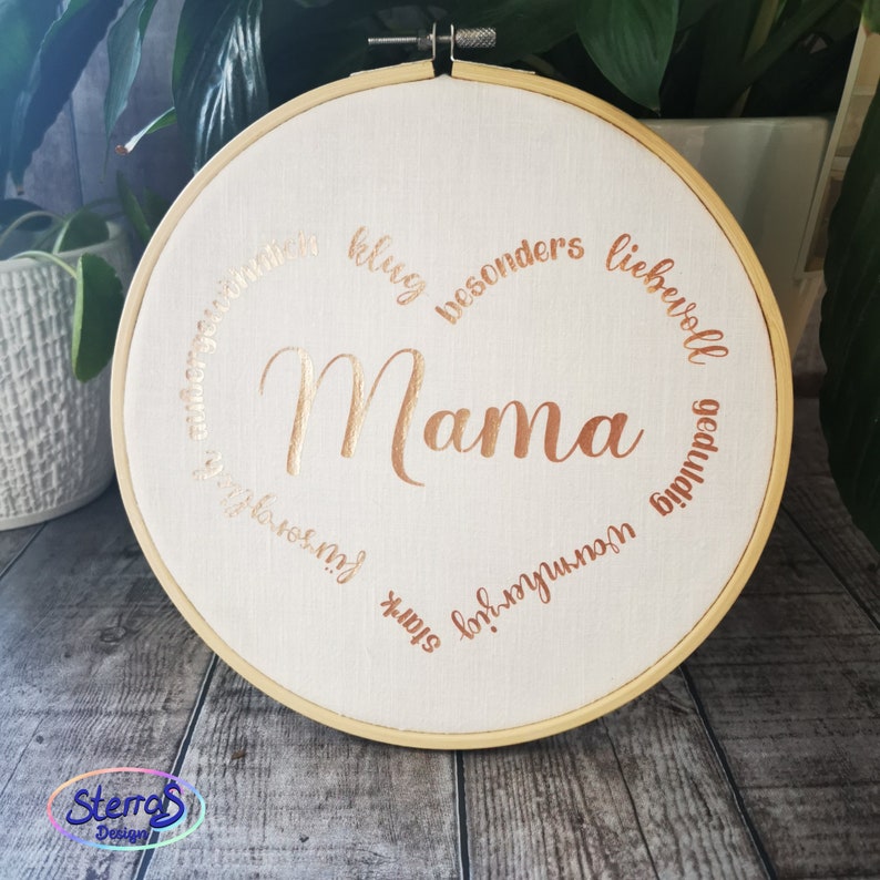 May include: A white embroidery hoop with a gold metallic inscription that reads "Mama" surrounded by a circle of text in German that translates to "loving, strong, unique, special, kind, patient, wonderful".