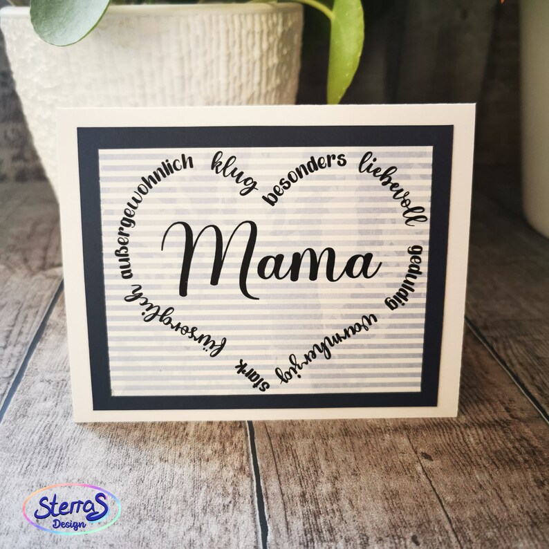 May include: A white card with a black border and a striped background. The word "Mama" is written in large, black, cursive letters. A circle of words surrounds the word "Mama" including "&uuml;bergew&ouml;hnlich", "klug", "besonders", "liebenswert", "geduldig", "stark", "toll", "wunderbar", "fantastisch", "f&uuml;rsorglich", and "au&szlig;ergew&ouml;hnlich".