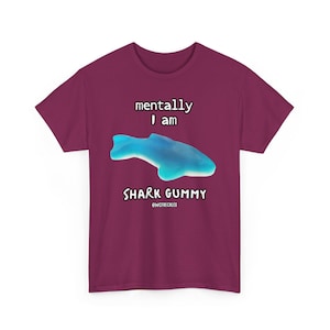 May include: A maroon t-shirt with the words "mentally I am SHARK GUMMY" and a blue gummy shark graphic. The text and graphic are centered on the front of the shirt. The shirt also includes the text "@WCFRECKLES" below the words "SHARK GUMMY".