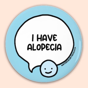 May include: A round, light blue button with the text "I HAVE ALOPECIA" in black within a white speech bubble. A small, smiling face is below the bubble. The button also has the text "WATERCOLOR FRECKLES" in black.