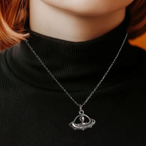 Könnte beinhalten: Eine silberfarbene Halskette mit einem Anhänger in Form eines UFOs, das eine Alienfigur und die Worte "I WANT TO BELIEVE" zeigt. Die Halskette wird über einem schwarzen Rollkragenpullover getragen. Der Anhänger hat einen Durchmesser von ca. 2,5 cm.
