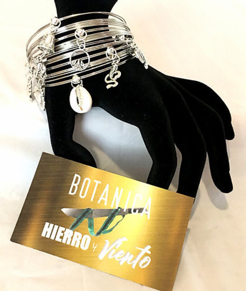May include: Silver charm bracelet with multiple charms, including a peace sign, a seashell, and a snake. The bracelet is on a black display stand with a gold tag that reads "BOTANICA HIERRO Y VIENTO".