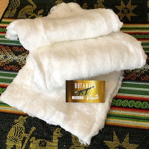 May include: A white, fluffy, cotton fabric folded in thirds. The fabric is on a colorful woven background with a gold label that reads "BOTANICA  HIERRO + VIENTO".