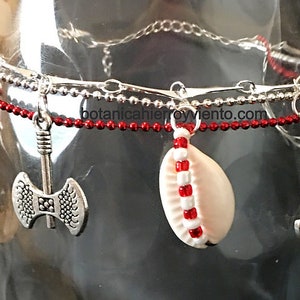 May include: A silver charm bracelet with red beads, a white seashell charm, and silver charms shaped like a sword, an axe, and a small axe. The bracelet is on a glass surface.