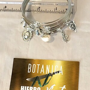 May include: A set of silver bangles with charms, including a pearl, a flower, and a coin with a design. The bangles are stacked on top of each other and are sitting on a ruler. The ruler is marked with inches. The image also includes a business card with the text "BOTANICA AL HIERRO Y VIENTO".