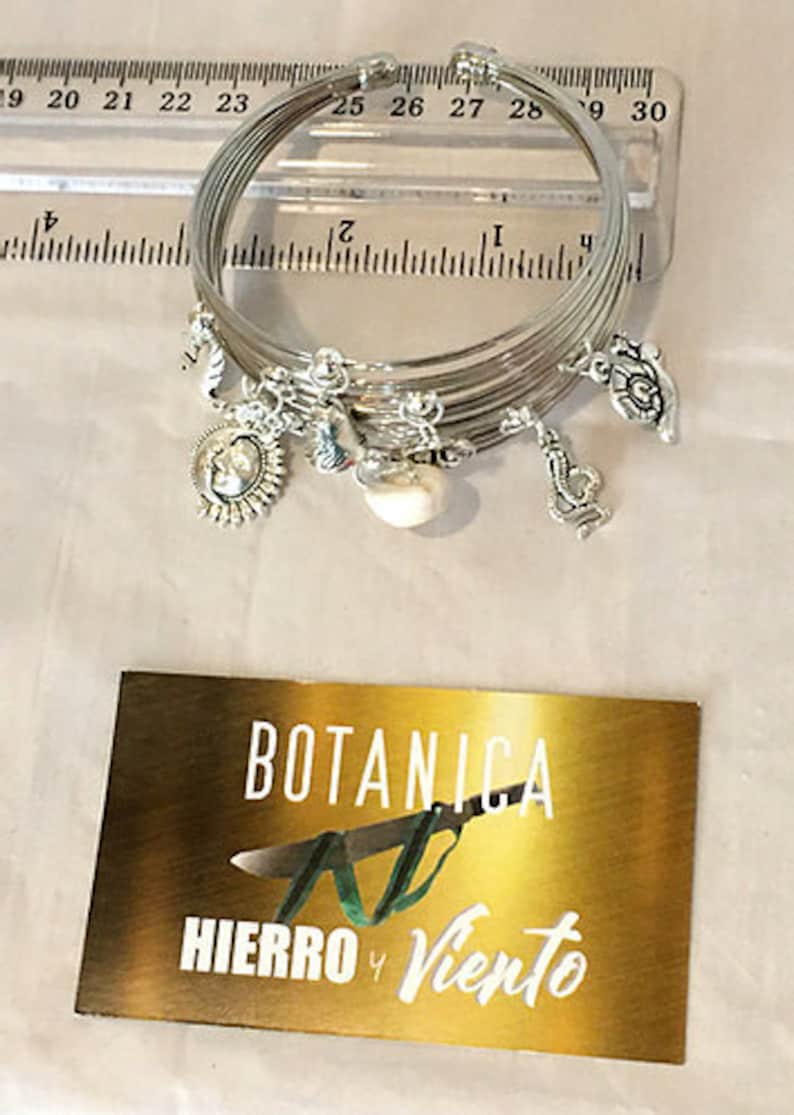 May include: A set of silver bangles with charms, including a pearl, a flower, and a coin with a design. The bangles are stacked on top of each other and are sitting on a ruler. The ruler is marked with inches. The image also includes a business card with the text "BOTANICA AL HIERRO Y VIENTO".