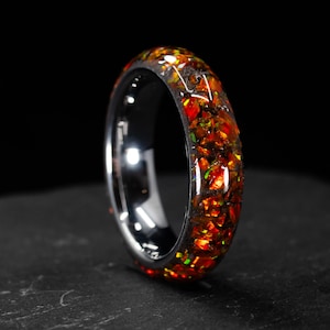May include: A close-up of a ring with a black interior and a band filled with fiery orange, red, and green fragments. The ring has a polished, silver-coloured exterior. The background is black.