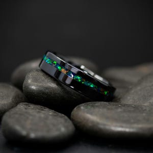 May include: A black ring with a vibrant green and blue opal inlay. The ring is set on a bed of dark gray stones, creating a striking contrast. The opal's colors shimmer, adding a touch of elegance.