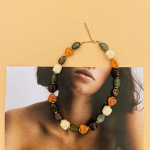 Peut inclure: Un collier composé d'une série de perles lisses et polies dans des tons vert foncé, crème, ambre et brun profond. Les perles sont entrecoupées de petites entretoises dorées et incluent des éléments en forme de cœur, avec une chaîne dorée.