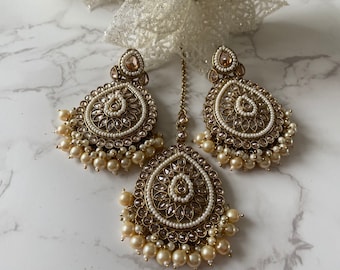May include: A set of gold-tone earrings and a matching maang tikka, featuring a teardrop design with white pearls and clear rhinestones.