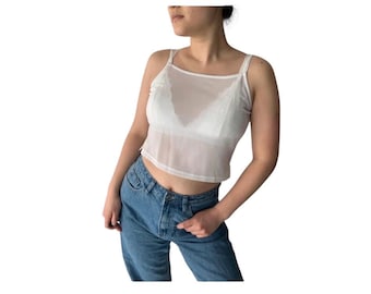 May include: A white sheer crop top with thin straps and a square neckline. The top is layered over a white bra. The model is wearing blue jeans.