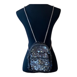 Puede incluir: Una pequeña mochila negra con lentejuelas y correas de cadena plateadas. La mochila tiene un compartimento principal con cremallera y un bolsillo frontal más pequeño. Las lentejuelas son iridiscentes, reflejando la luz. La mochila se muestra en un maniquí negro.