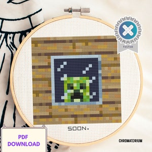 May include: A cross-stitch pattern of a green Minecraft Creeper in a wooden frame. The pattern is in a hoop and the text "SOON." is below the hoop.
