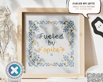 May include: A framed cross-stitch pattern with the text "Fueled by Spite" in green and yellow. The design is surrounded by blue and yellow floral accents. The text "Fueled by Spite" is in a playful font. The frame is light wood.