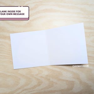 May include: A blank white card with a folded edge. The card is on a light brown wooden surface. The text "BLANK INSIDE FOR YOUR OWN MESSAGE" is on a pink rectangle in the top left corner.