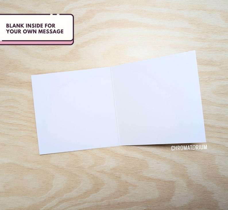 May include: A blank white card with a folded edge. The card is on a light brown wooden surface. The text "BLANK INSIDE FOR YOUR OWN MESSAGE" is on a pink rectangle in the top left corner.